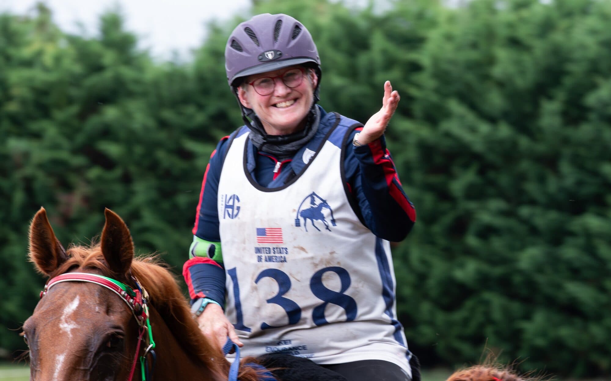Holly Corcoran on a chestnut horse, wearing a United States of America rider bib, waving.